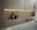 Recessed shelf in a gray tiled bathroom holding three bath products with warm backlighting above a white bathtub and a metal faucet to the right